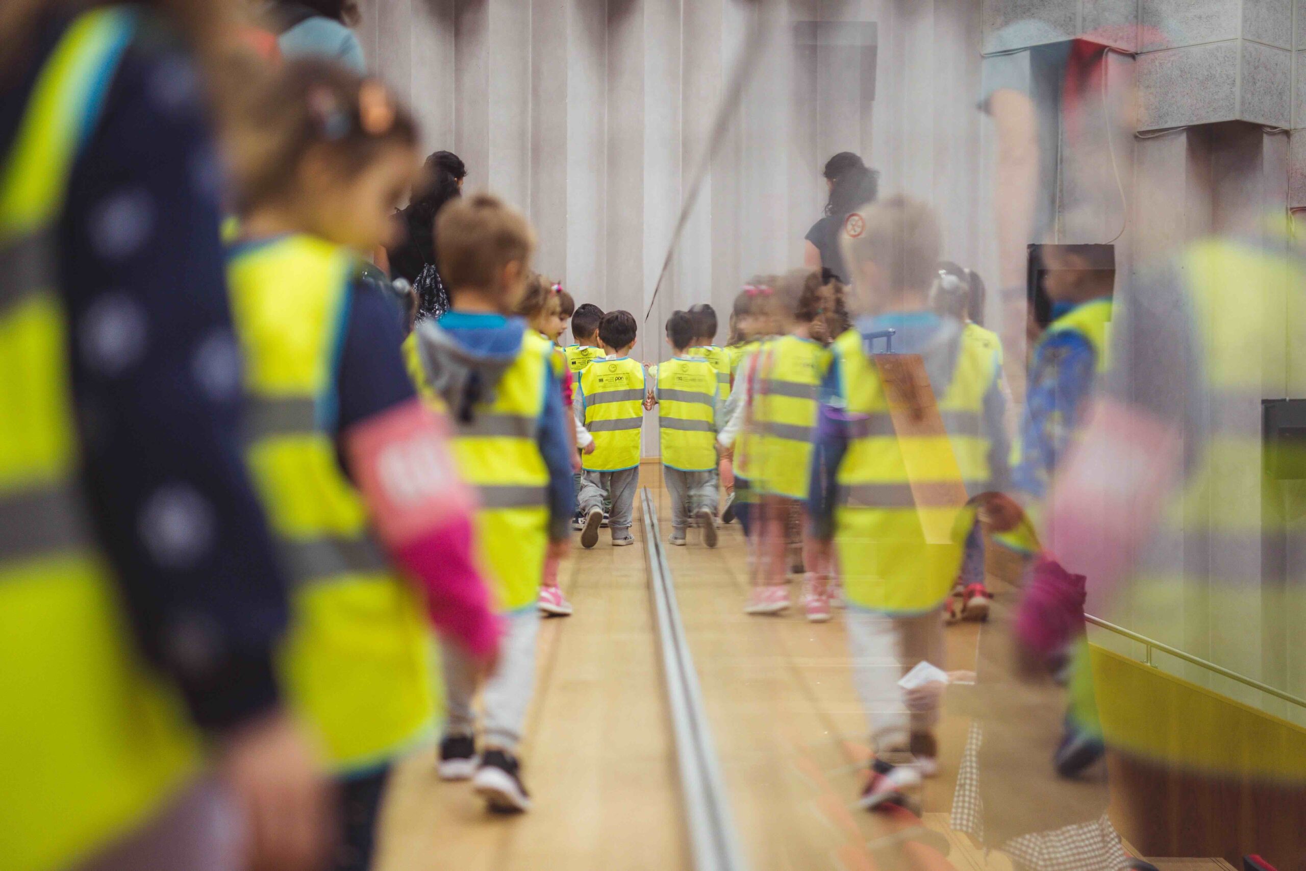 bambini con giubbini color giallo camminano riflessi nel vetro di protezione a teatro