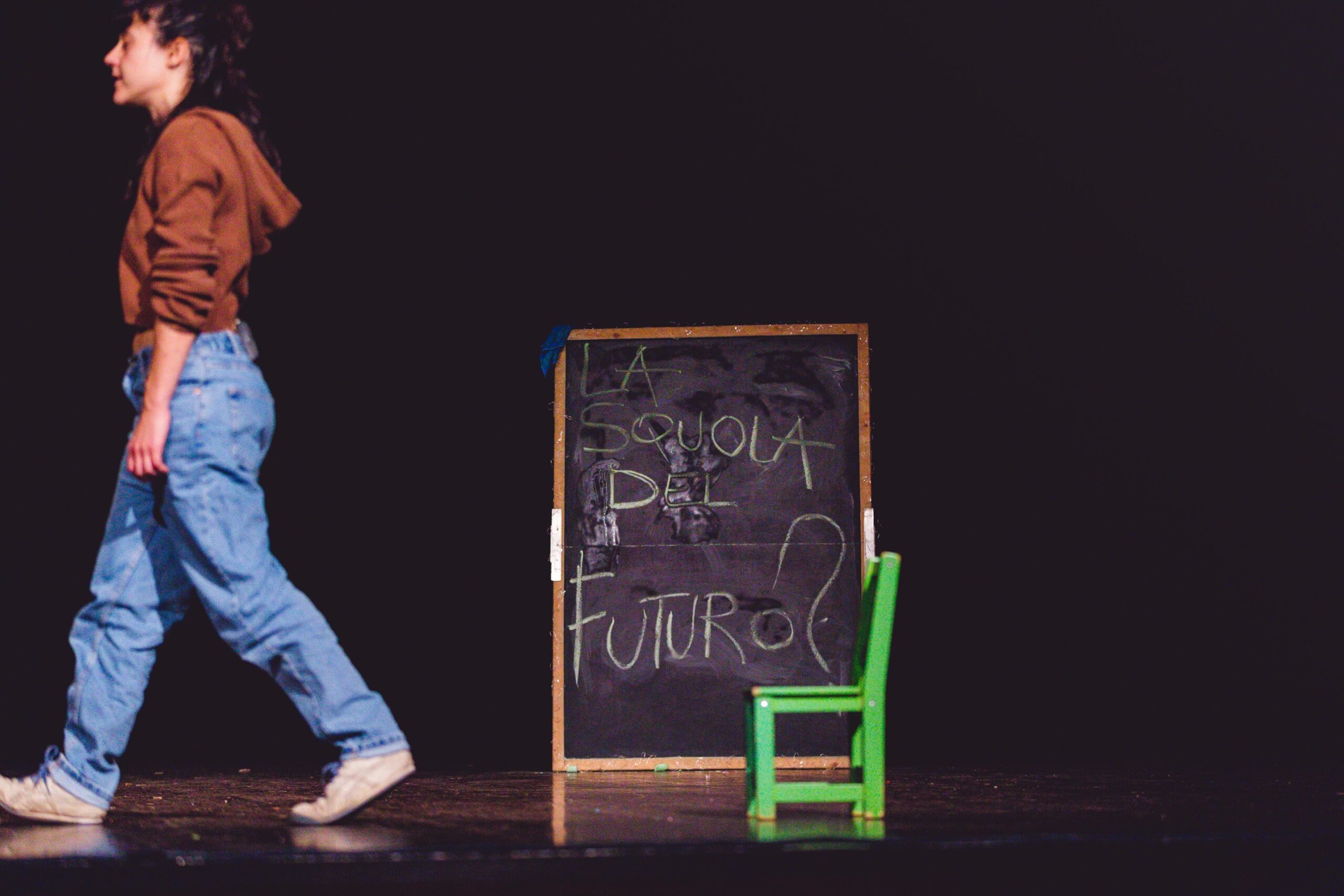 un'attrice che cammina sul palco con alle spalle una lavagna da scuola con la scritta La scuola del futuro?